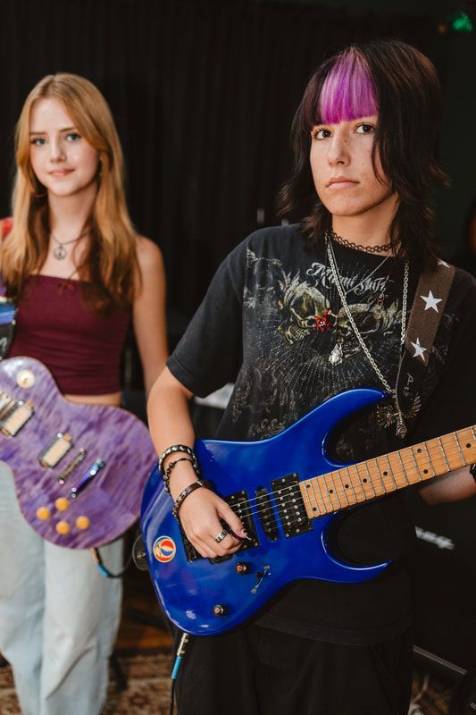 A girl with purple hair is holding a blue guitar next to another girl holding a purple guitar.
