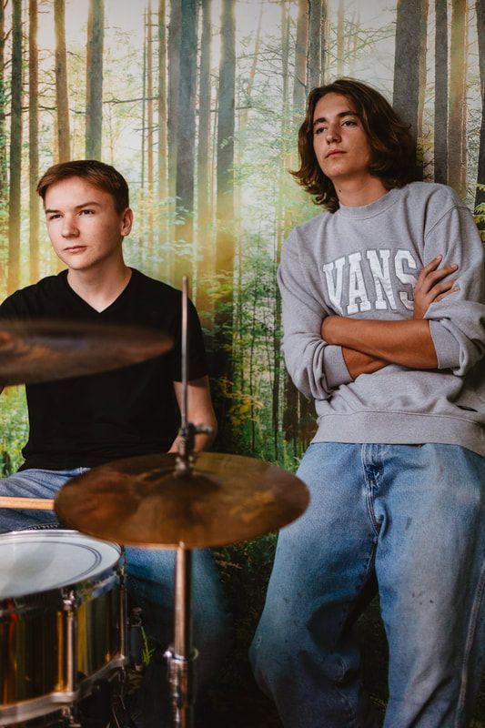 Two young men are standing next to each other in front of a drum set.