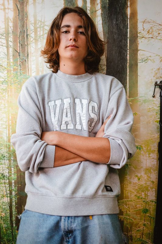 A young man wearing a grey vans sweatshirt is standing with his arms crossed.