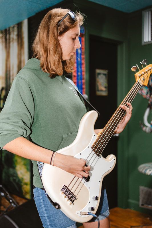 A woman in a green hoodie is playing a white bass guitar
