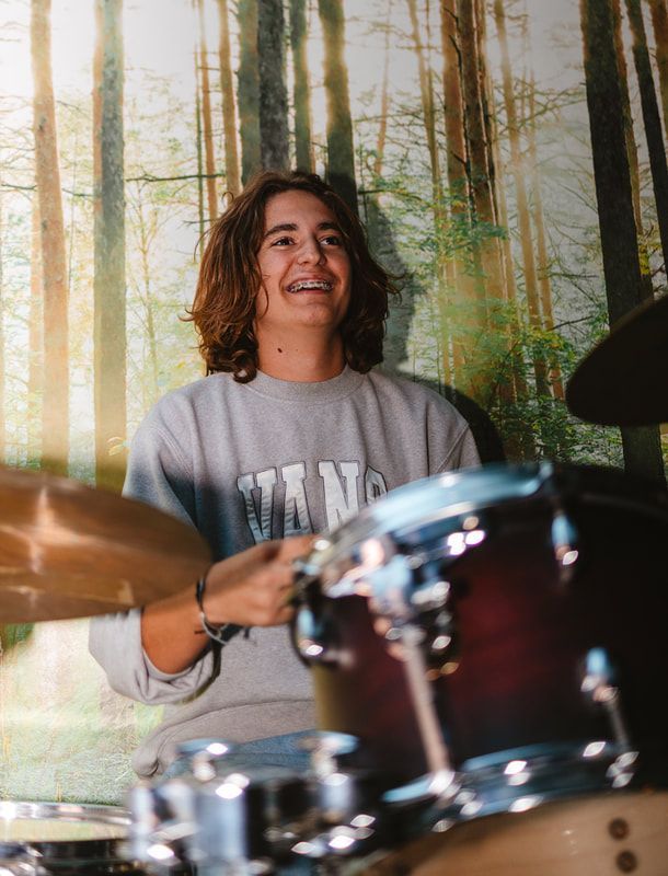 A young man is playing drums in front of a forest background.