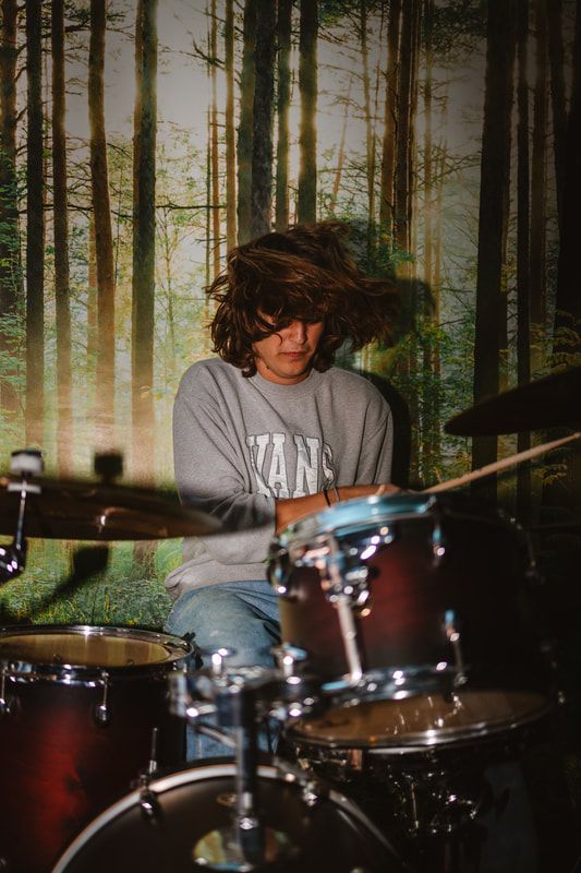 A man is playing drums in front of a forest background.
