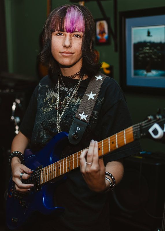 A young woman with purple hair is playing a blue electric guitar.