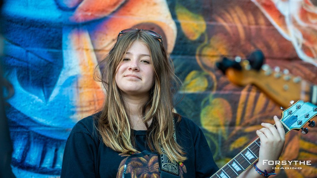 A young woman is holding a guitar in front of a graffiti wall.