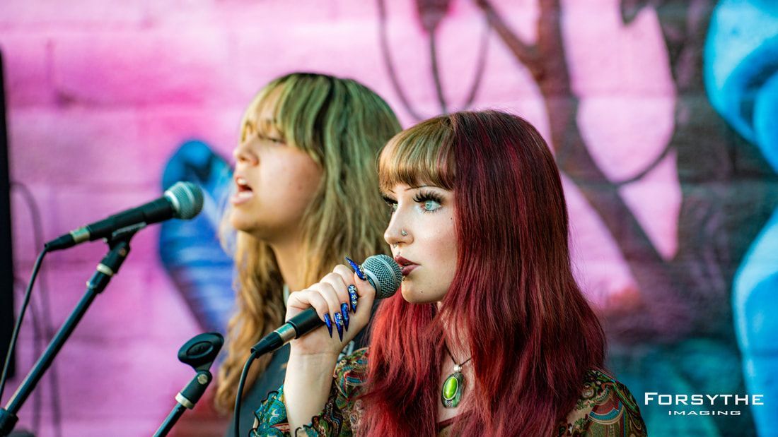 Two women are singing into microphones in front of a pink wall.