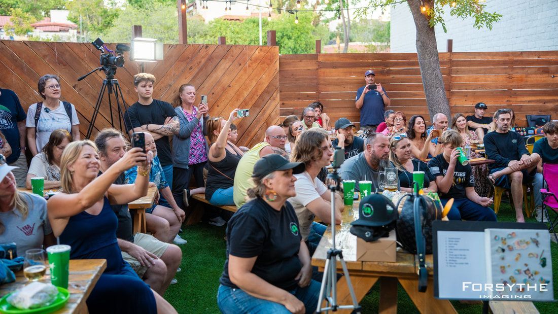 A large group of people are sitting at picnic tables in a backyard.