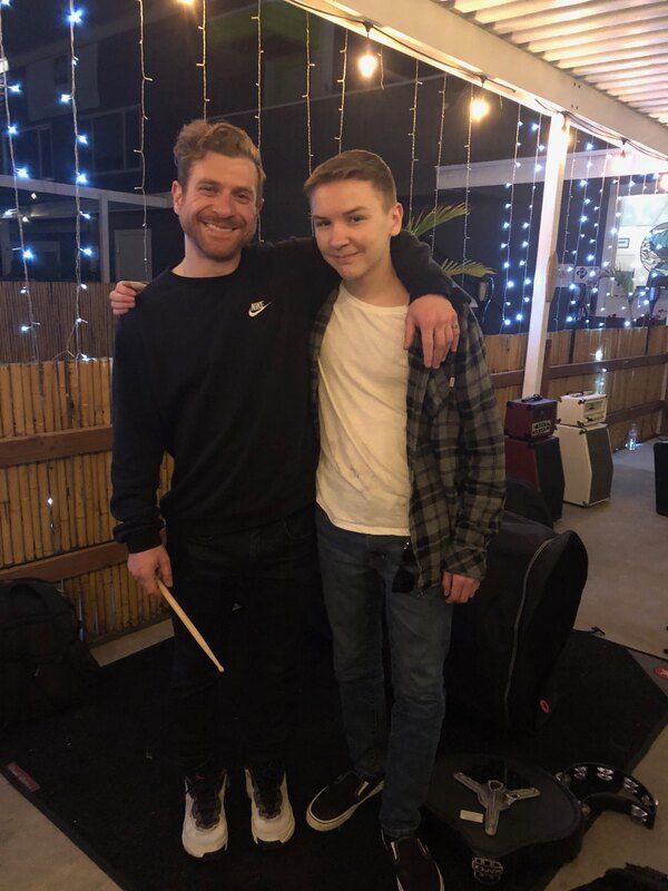 Two men are standing next to each other and one is holding a drum stick.