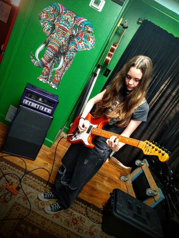 A girl is playing an electric guitar in front of a green wall with an elephant on it