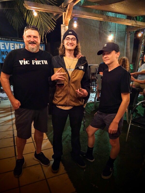 A man wearing a vic firth shirt is standing next to two other men