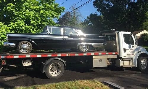 Black classic car on a flatbed tow truck. Sunny outdoor setting with green foliage.