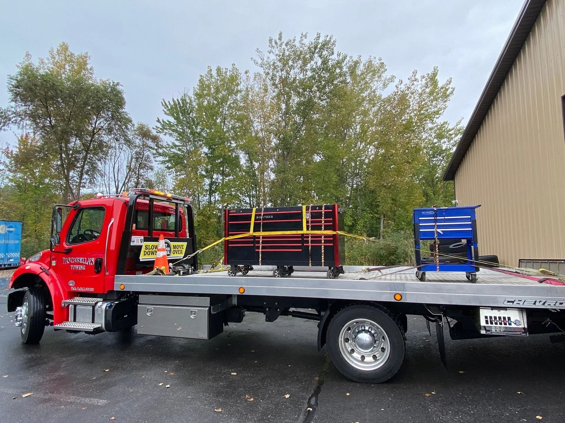 Red tow truck transporting two tool chests on a flatbed, yellow straps securing the cargo.