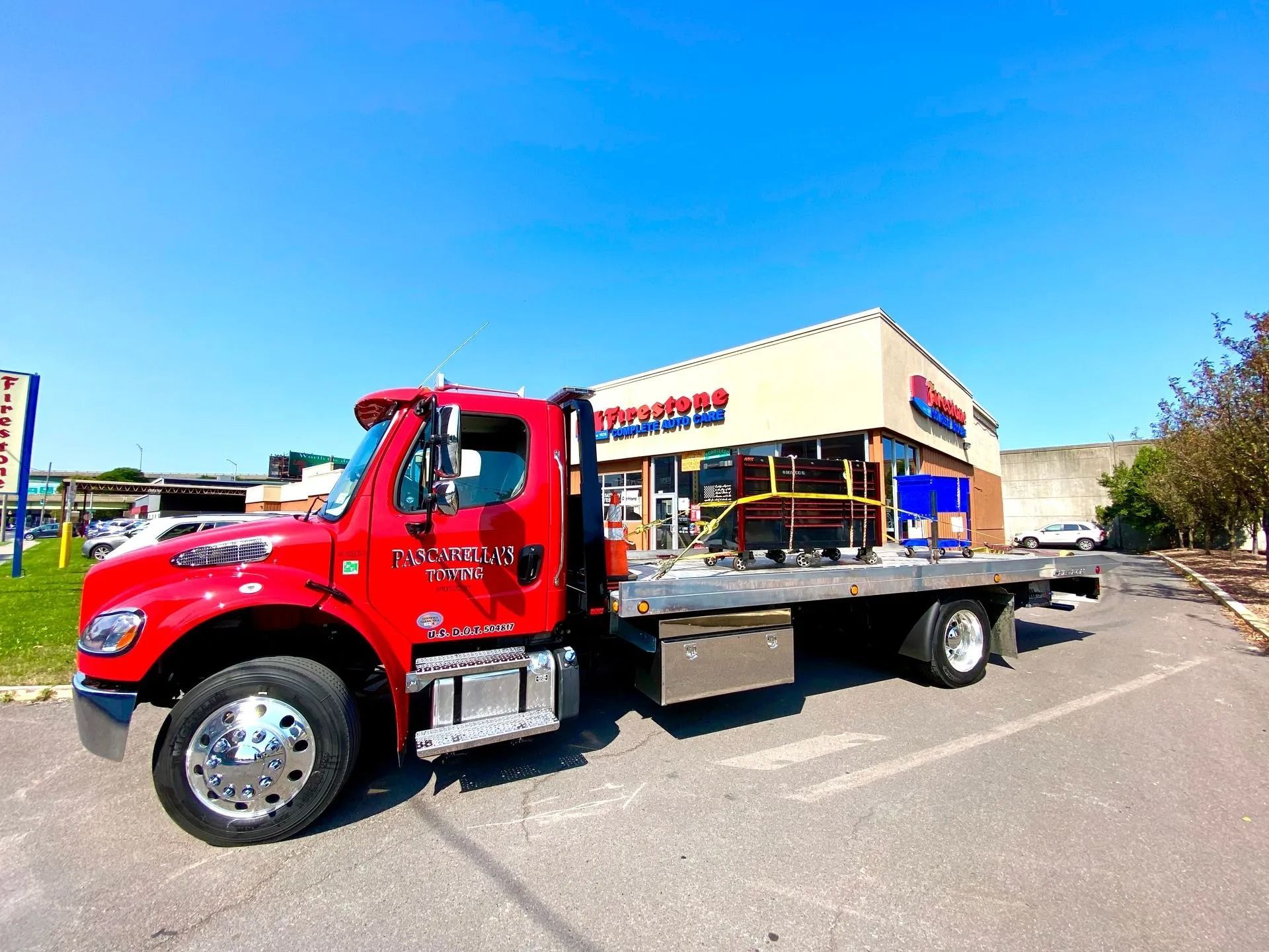 Red flatbed tow truck parked in front of a building with red and blue signage; clear blue sky.