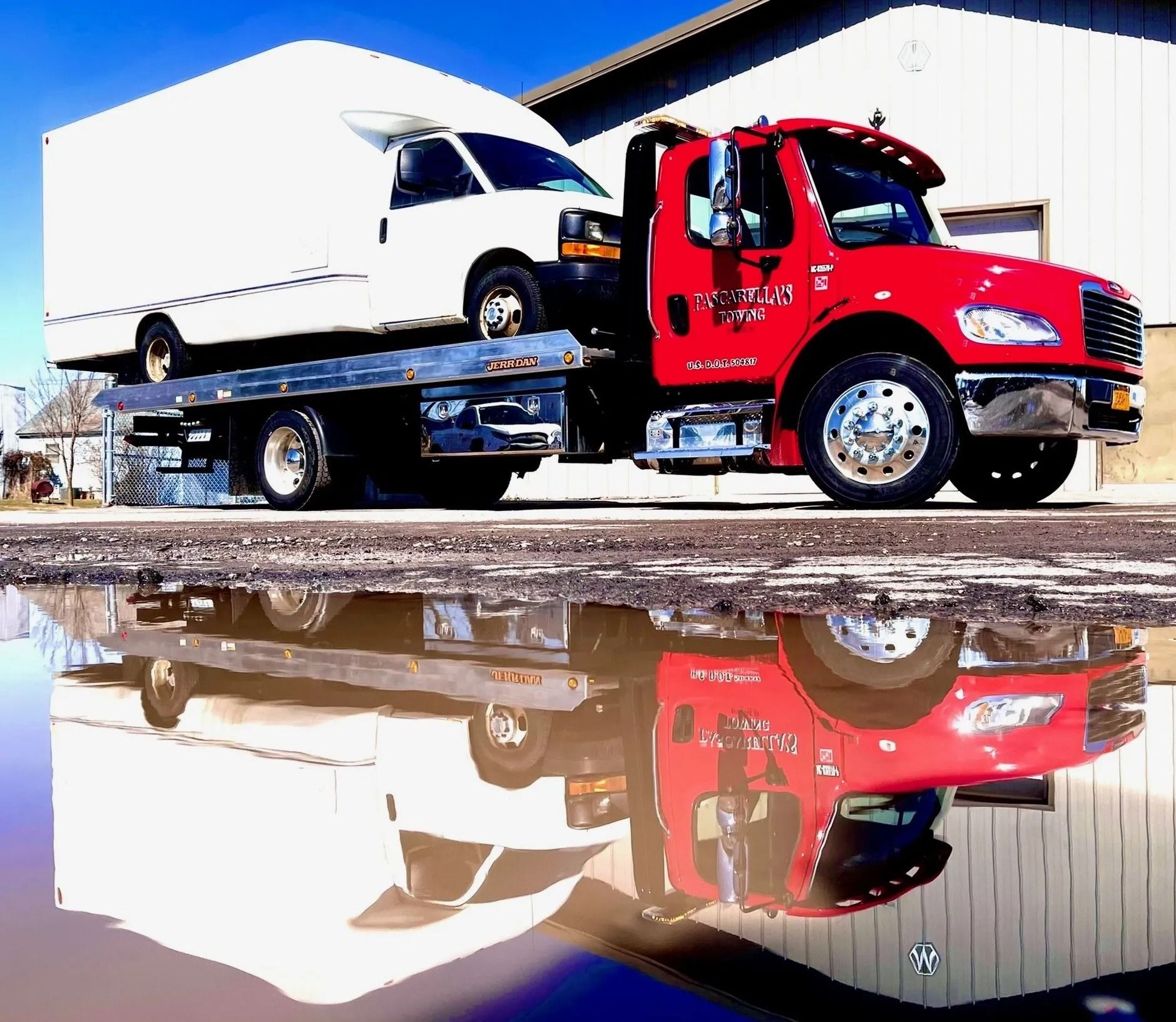 Red tow truck carrying a white van, reflected in a puddle.