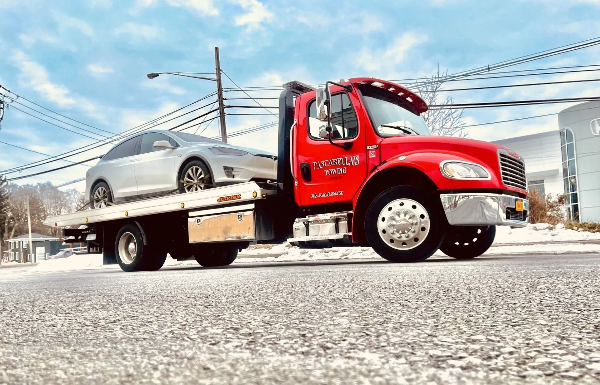 Red tow truck with a silver car on its flatbed, parked on a snowy road under a blue sky.