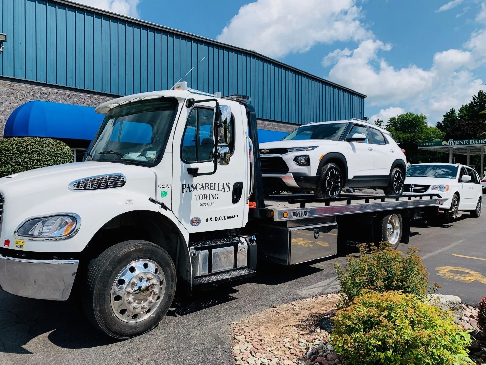 White tow truck carrying a white SUV and a white pickup truck, parked in front of a blue building.