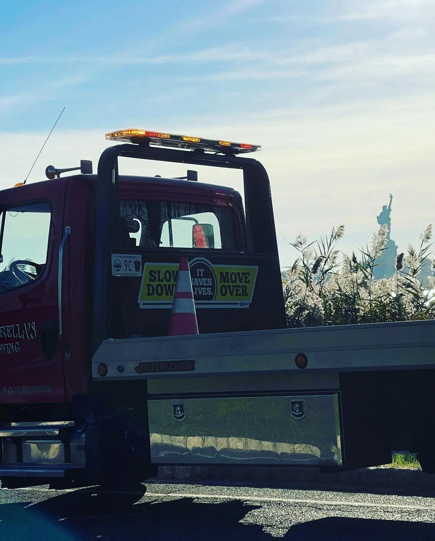Red tow truck with warning lights, cone, and “Slow Down Move Over” sign, Statue of Liberty in background.