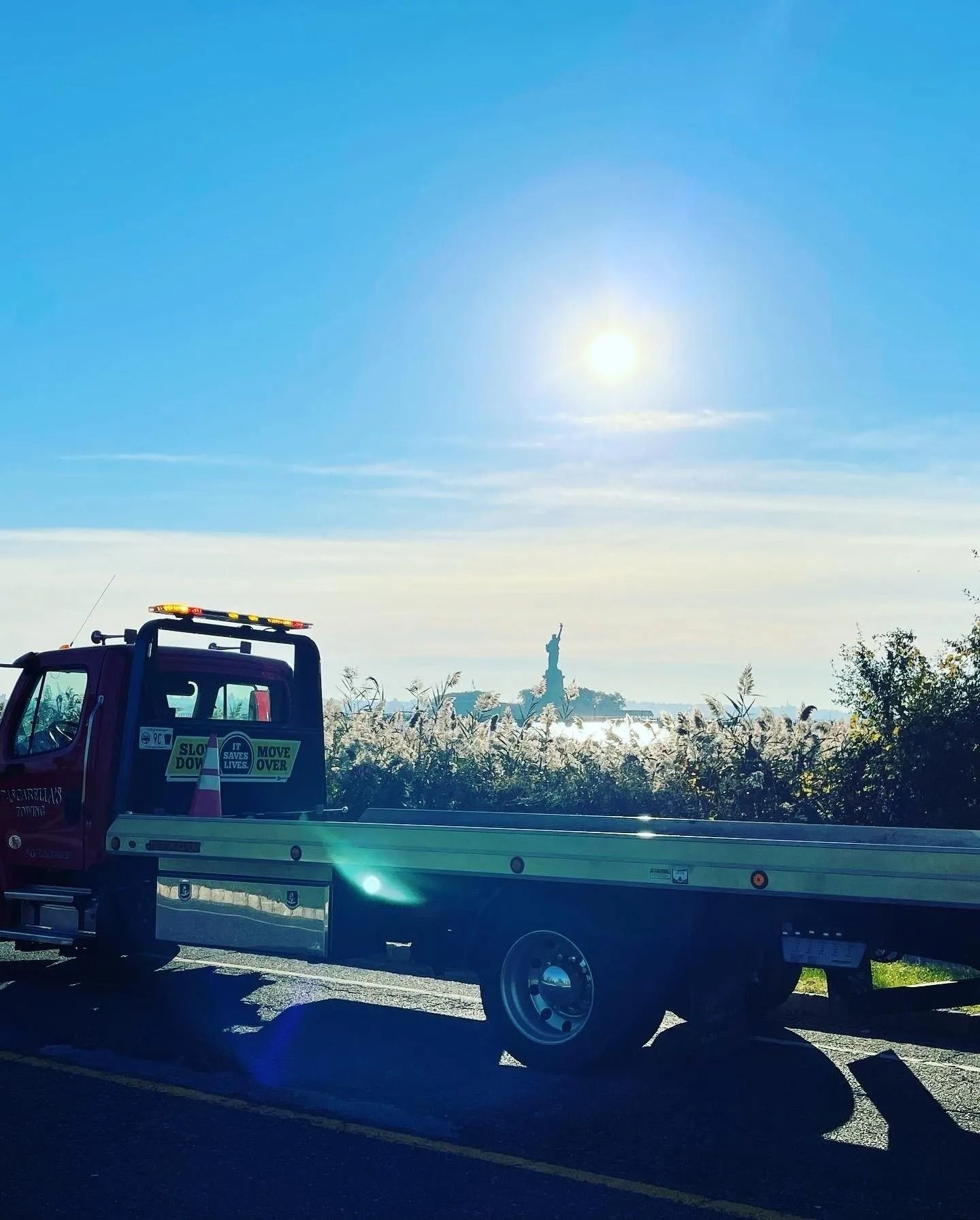 Tow truck parked with the Statue of Liberty visible in the distance under a sunny sky.