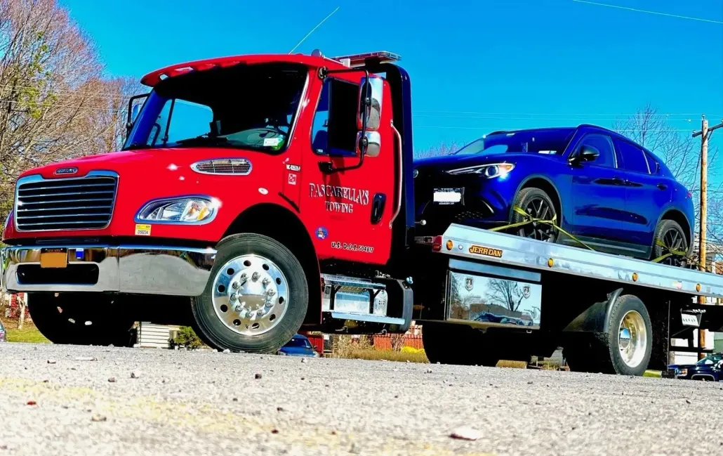 Red tow truck hauling a white box truck. The sky is blue and a white building is in the background.