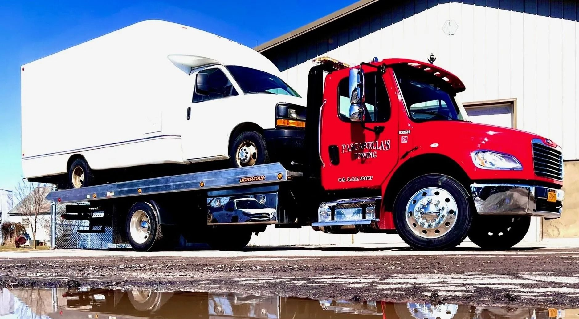 Red tow truck hauling a white box truck. The sky is blue and a white building is in the background.