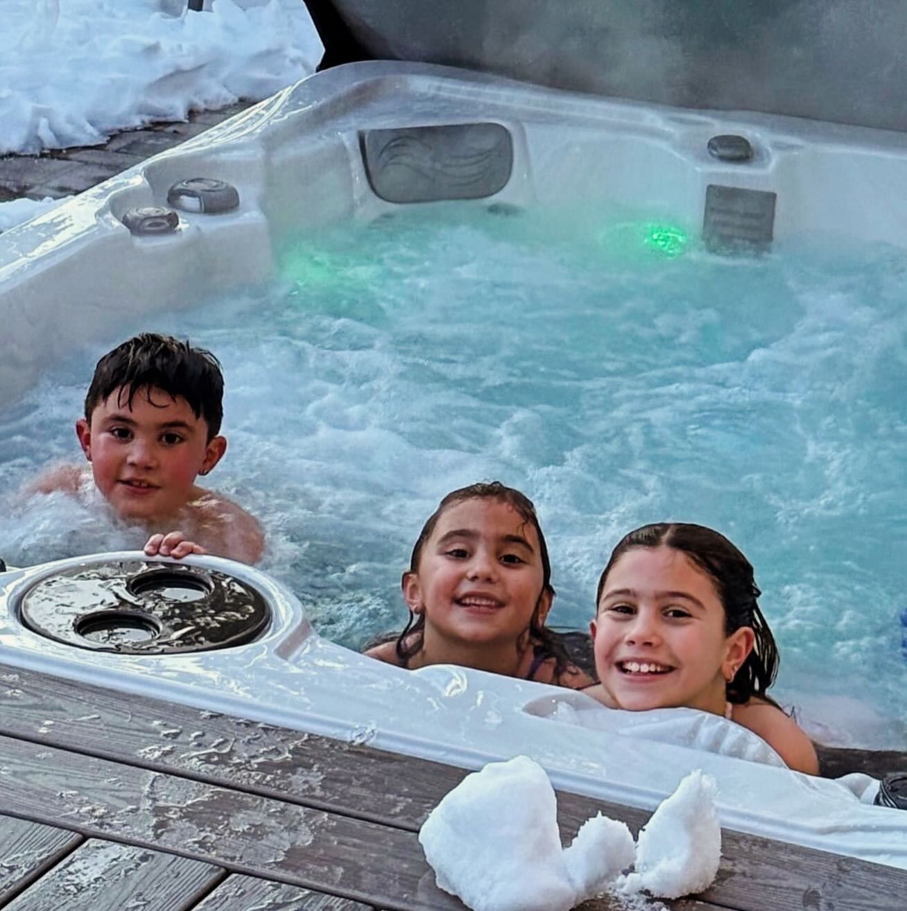 Three smiling children in a snowy hot tub with bubbling water and green lights.