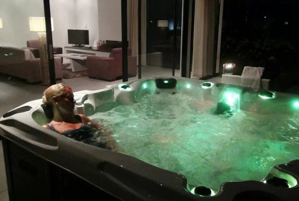 Person relaxing in a lit outdoor hot tub at night, with house lights visible nearby