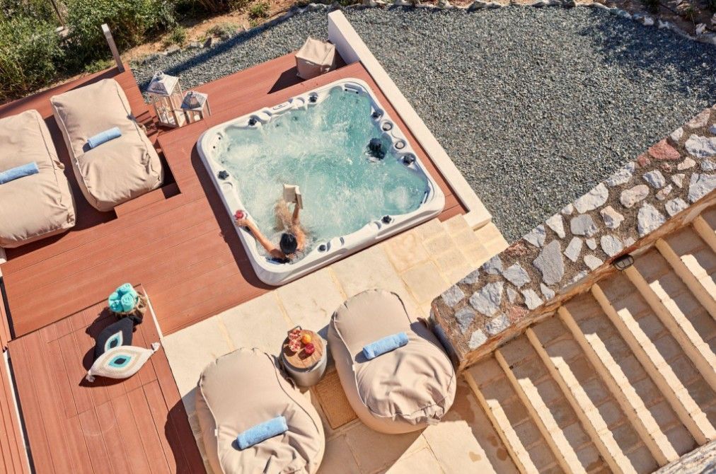 Aerial view of a hot tub on a wooden deck with cushions, a person relaxing inside, and stone landscaping nearby