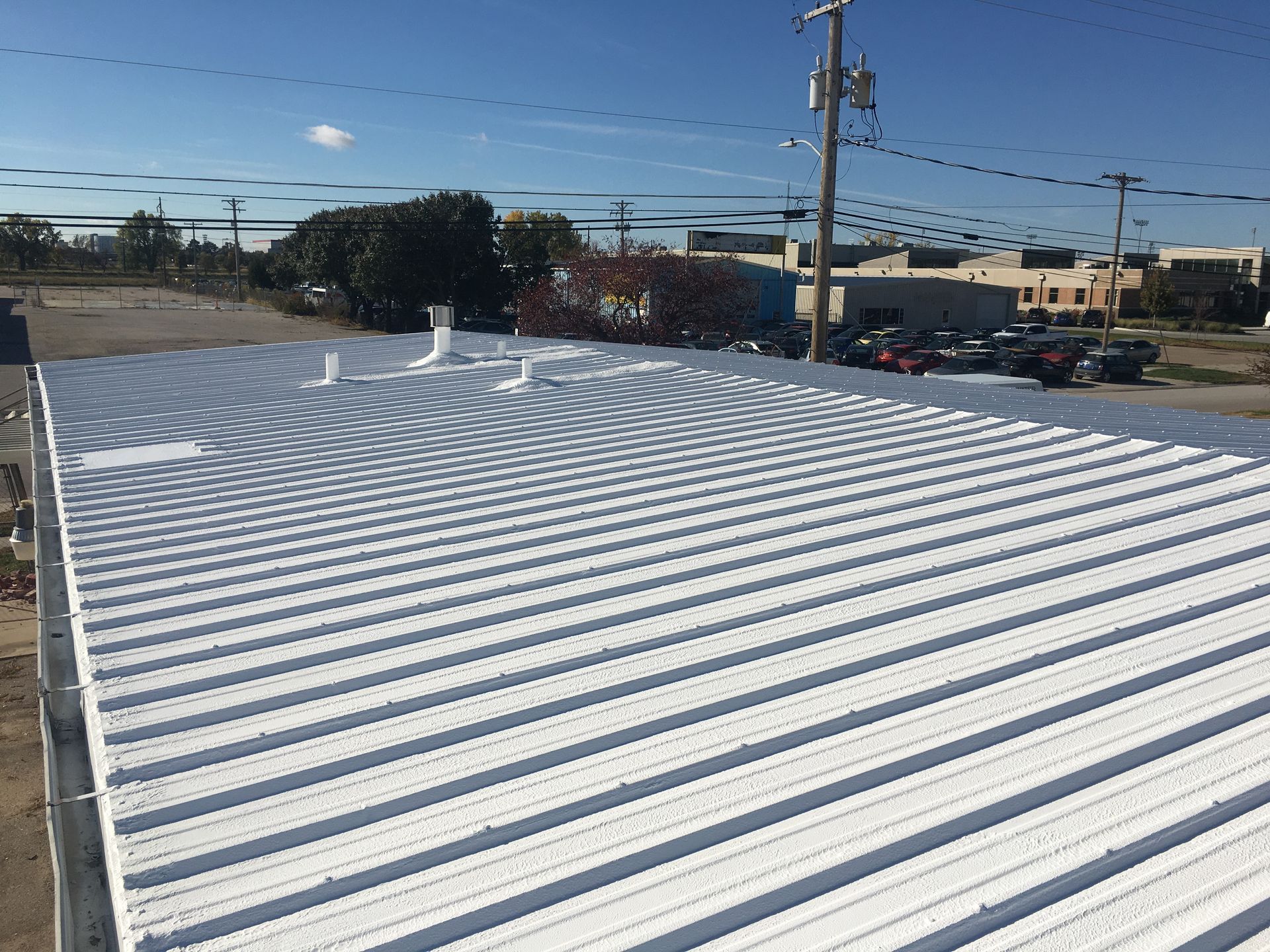 White corrugated metal roof on a building, under a bright blue sky with trees and buildings in background.