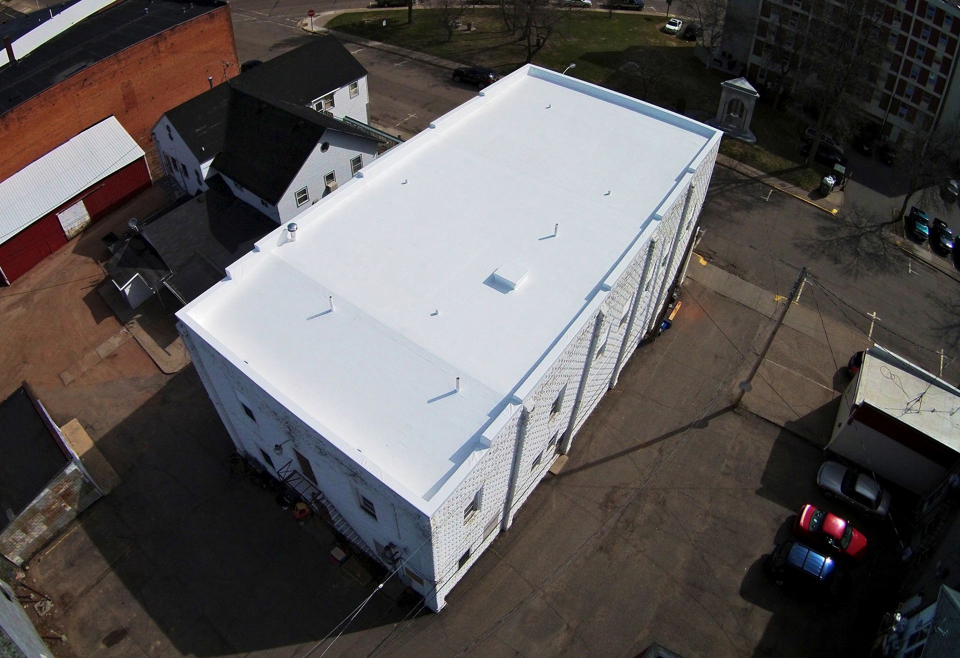 Aerial view of a white building with a flat, white roof in an urban setting. Sunlight casts shadows.