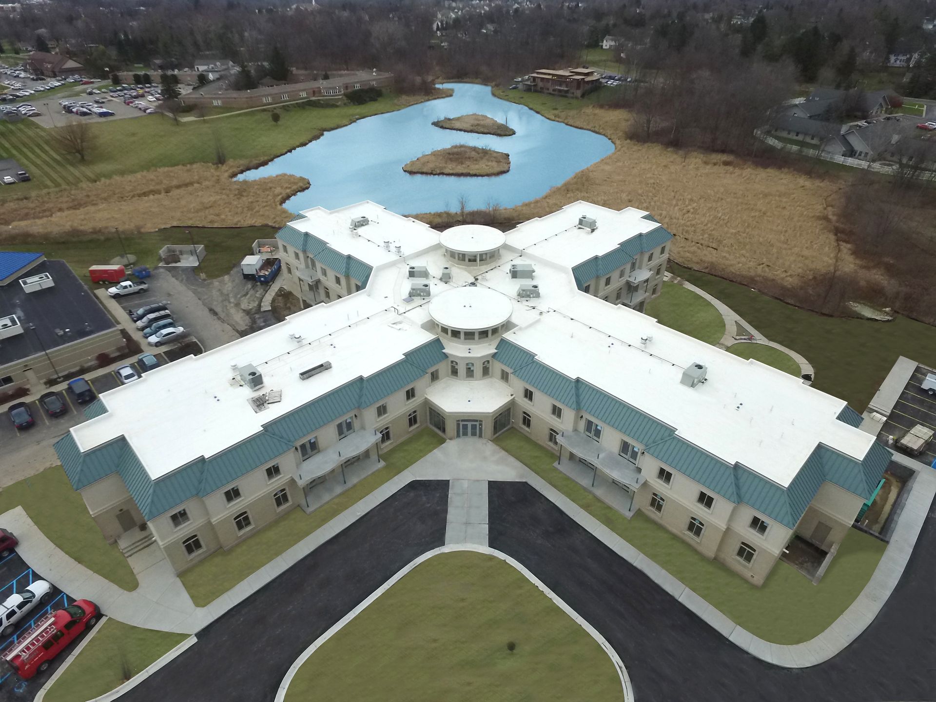 Aerial view of a cross-shaped building with a blue-roofed facade. A pond with islands is in the background.