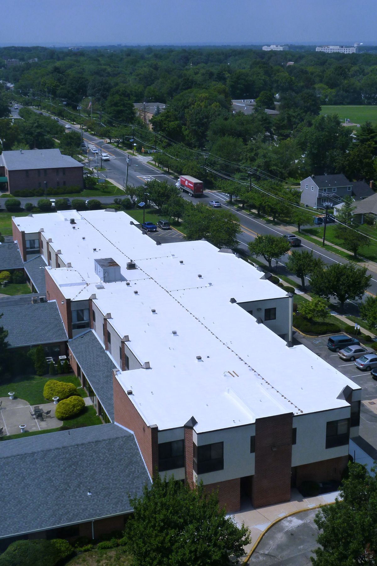 Aerial view of a long, white-roofed building with a brick facade, a road, and a tree-filled landscape in the background.