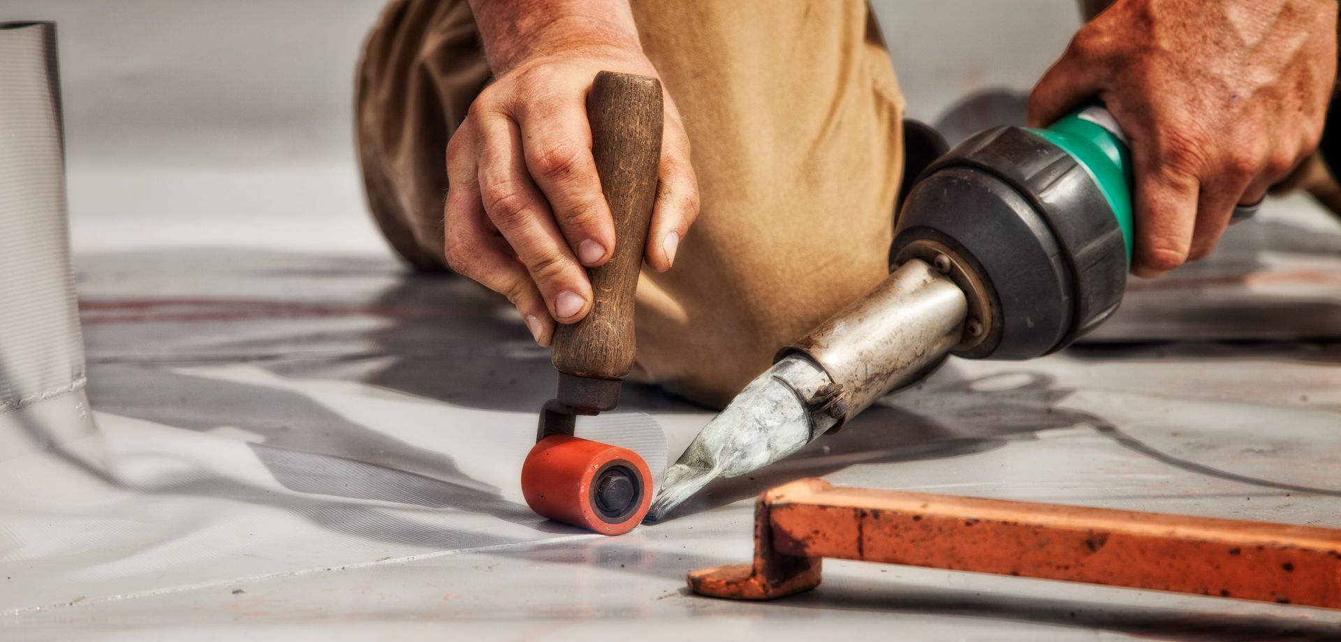Person uses a heat tool and roller to weld flooring seams on a gray surface. Brown pants, orange frame.