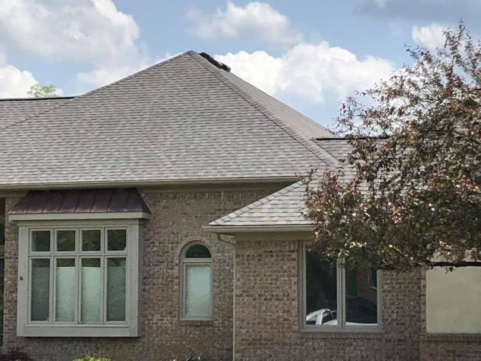 Brick house with gray shingle roof, bay window, and a tree with red leaves.