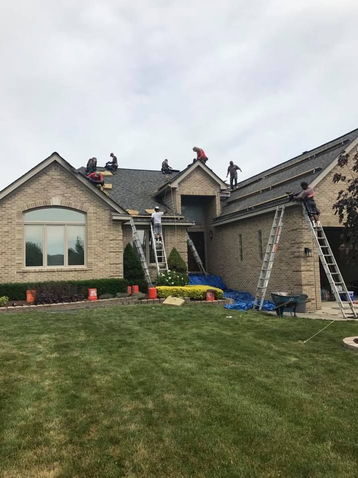 Roofers on a house with ladders and blue tarp, removing/replacing roof shingles under an overcast sky.
