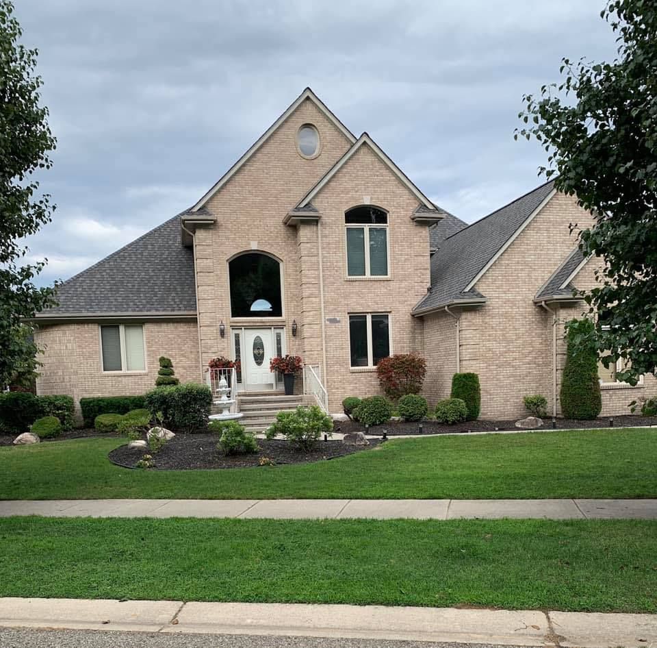 Two-story brick house with dark roof and well-manicured lawn and landscaping on a cloudy day.