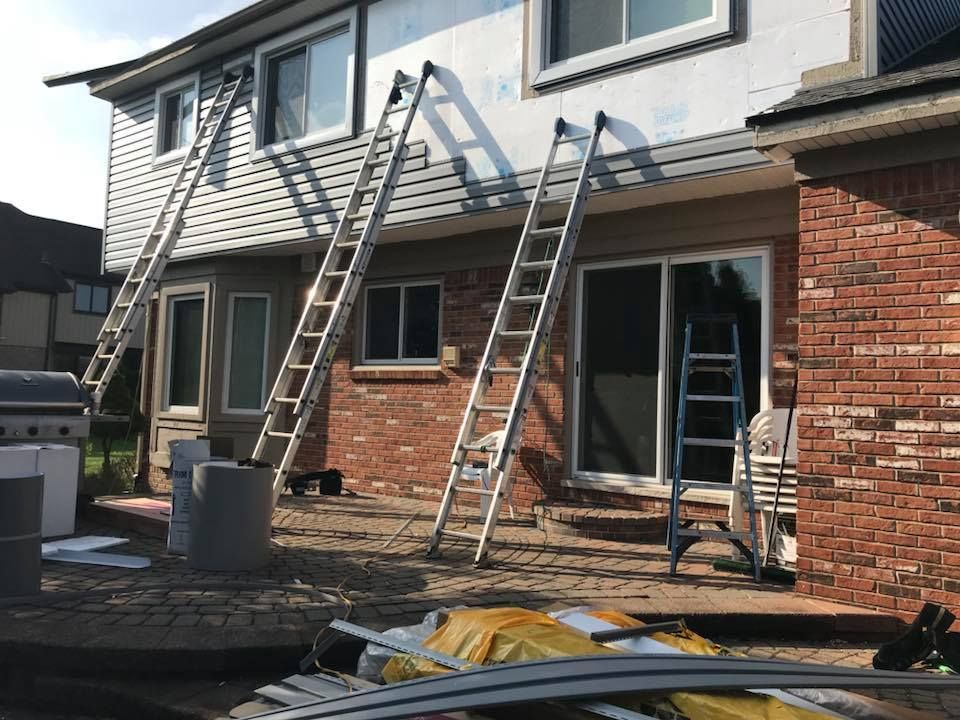 Ladders propped against a house with siding being installed. Brick and gray siding are visible.