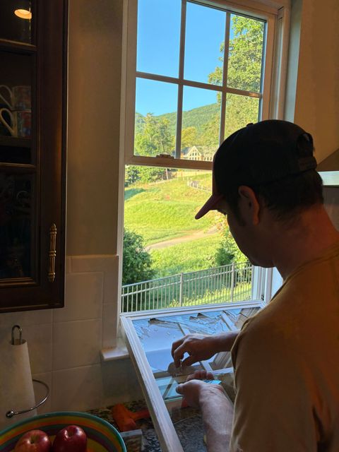 Person washing dishes at a kitchen sink by a bright window overlooking green hills.