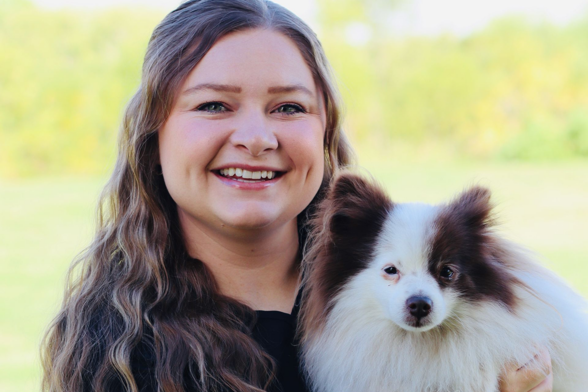 Woman smiles, holding a fluffy white and brown dog outdoors in front of greenery.