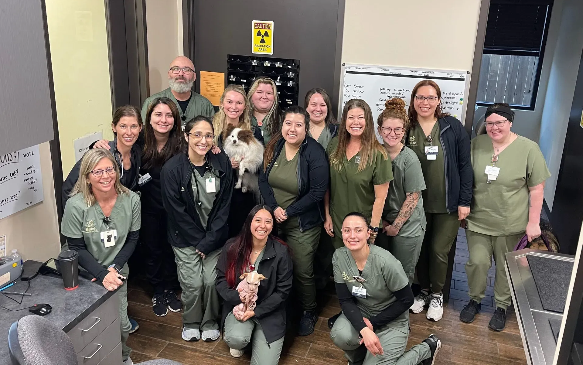 Veterinary clinic staff pose together. Several wearing green scrubs, one holding a small dog. Indoors with equipment.
