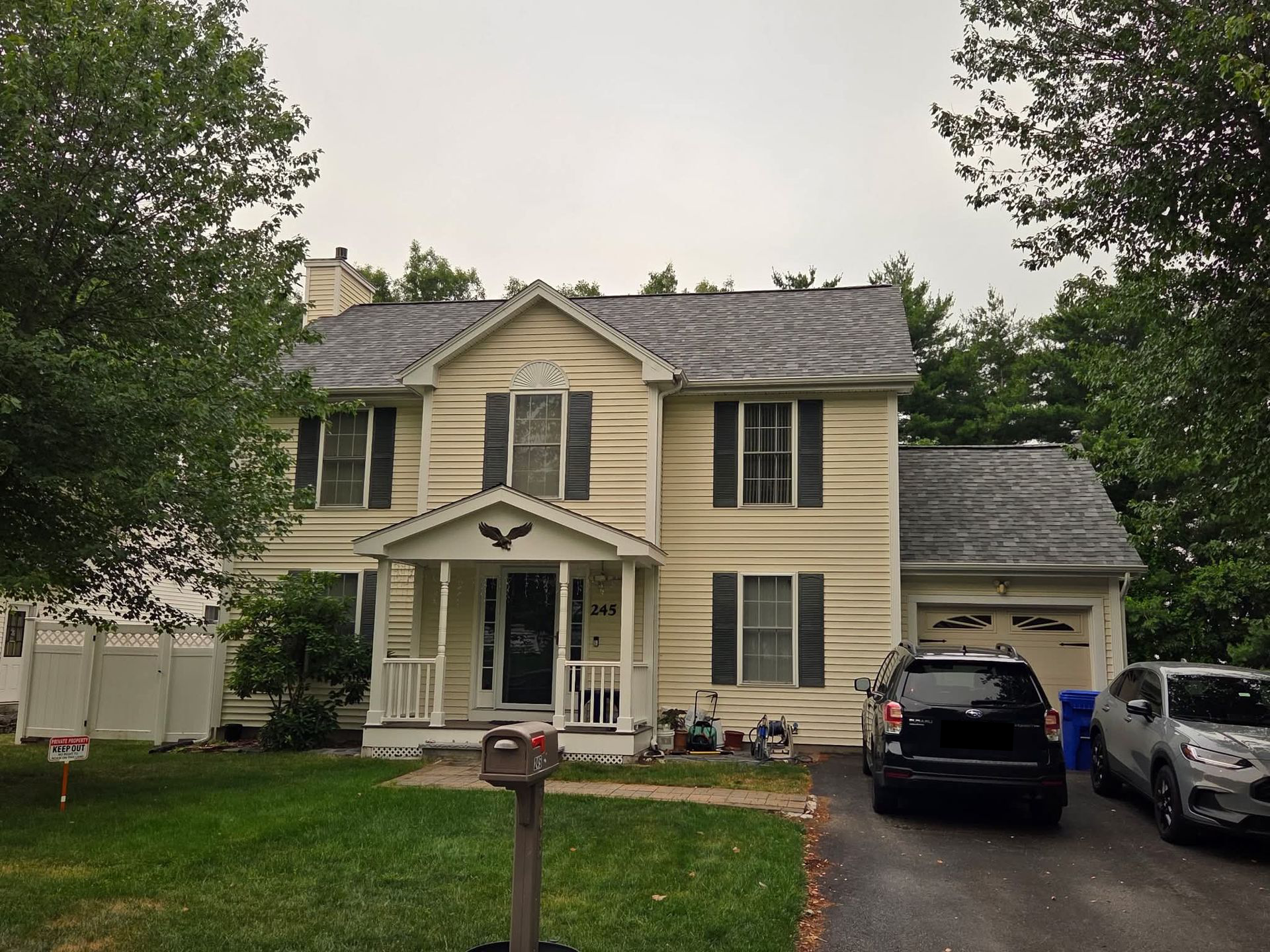 Two-story beige house with a front porch, black SUV in the driveway, and trees framing the yard.