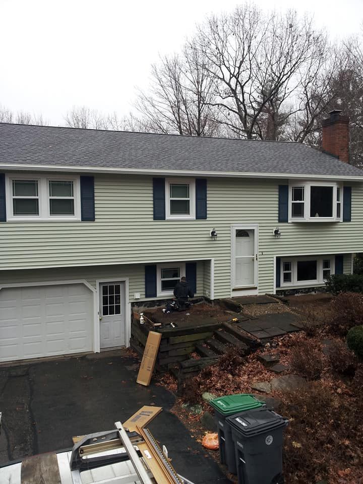 Two-story house with light green siding, white garage door, and dark blue shutters. Driveway leads to the house.