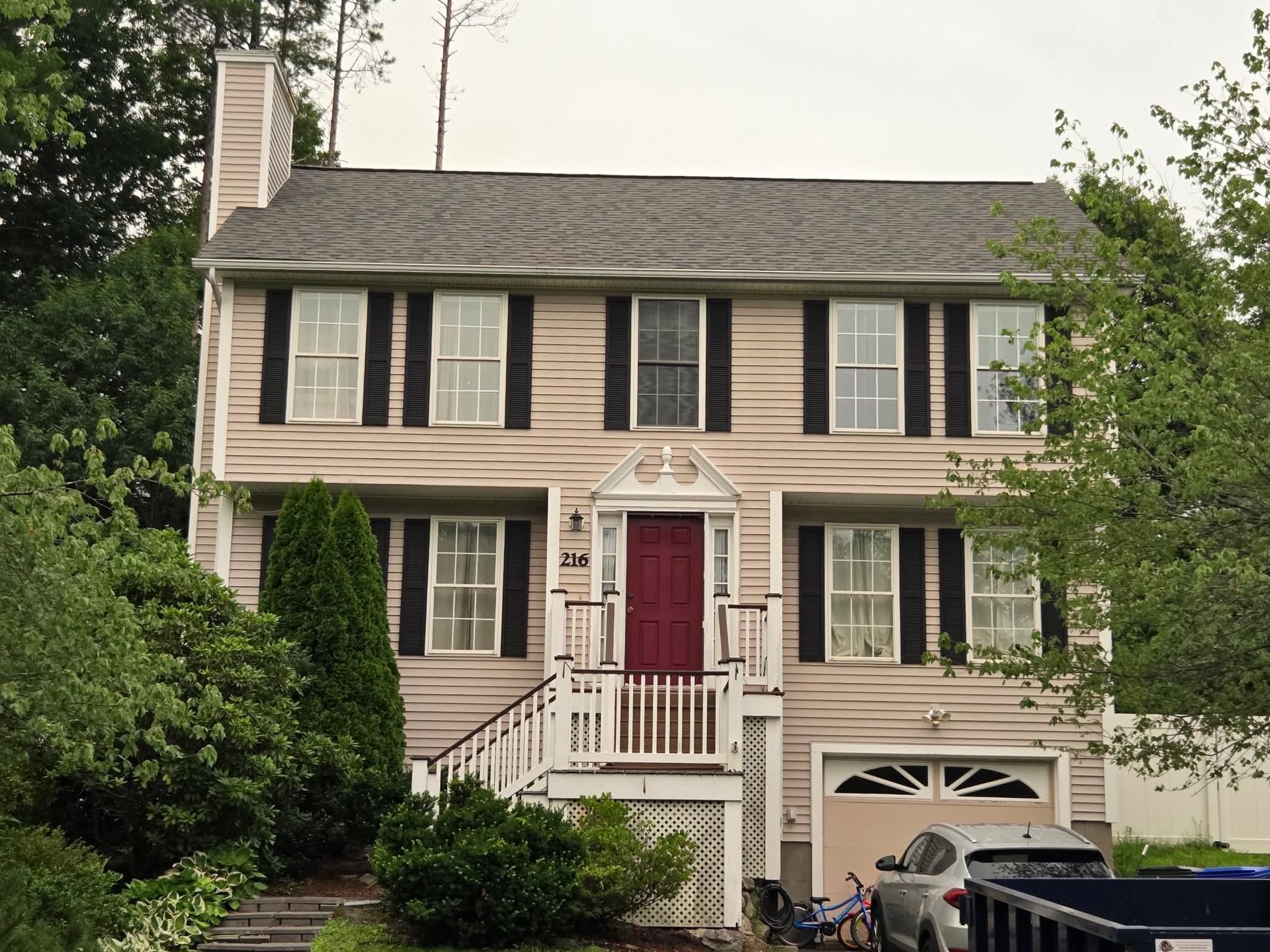 Two-story beige house with black shutters, a red front door, white porch, and a garage at the right.