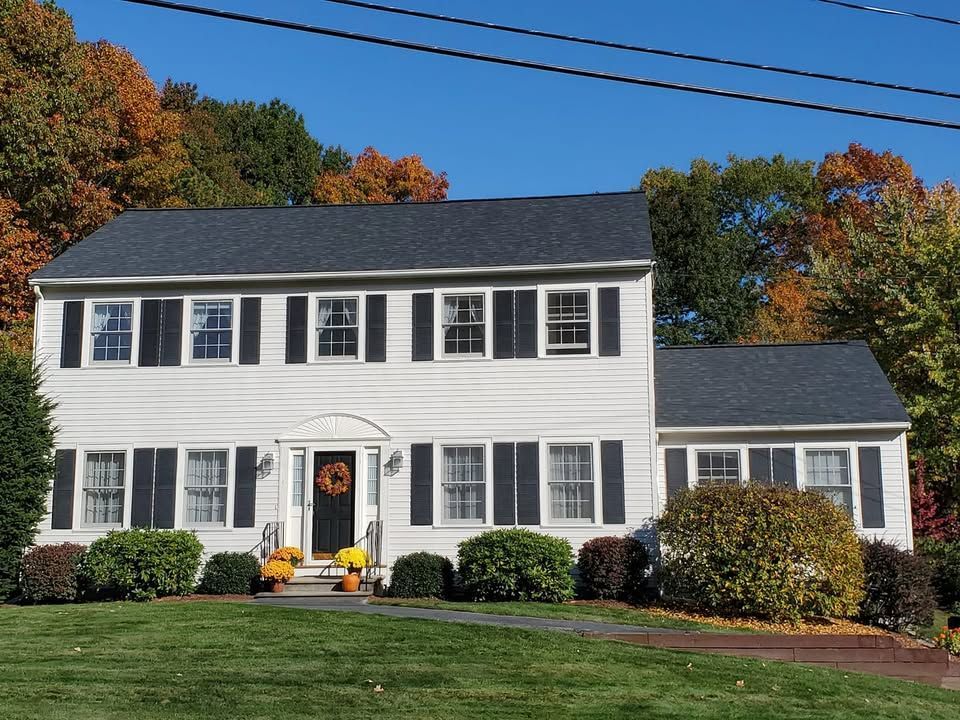 White two-story house with black shutters, black roof, and autumn foliage in background.