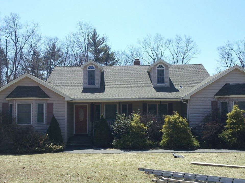 Light pink house with a gray roof and two dormer windows, surrounded by trees and bushes.
