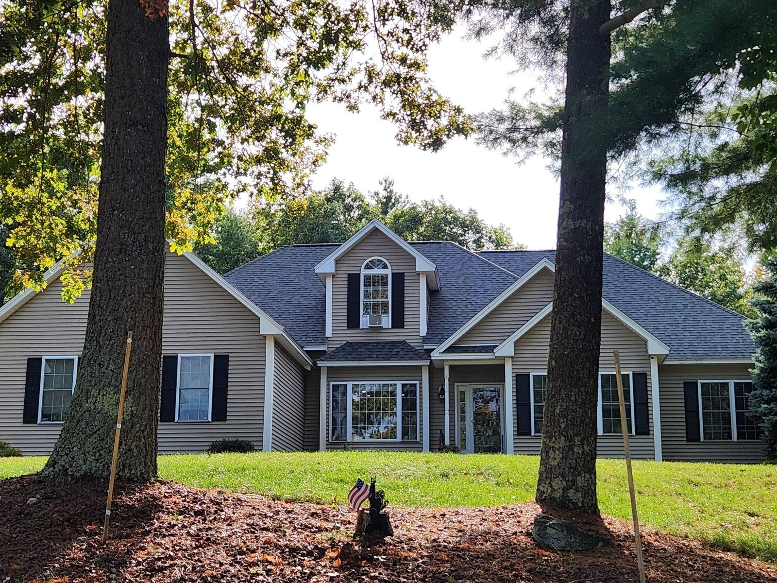 Tan house with black shutters, dormer, and gray roof, viewed through trees.