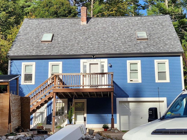 Two-story blue house with gray roof, deck, and garage. A white vehicle is partially in view.