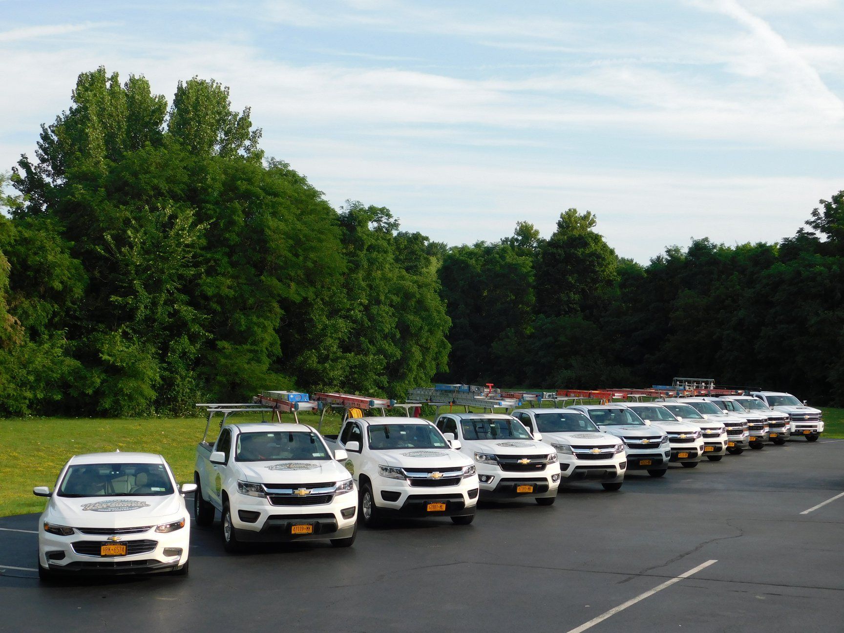 Row of white work trucks parked on asphalt in front of trees under a cloudy sky.