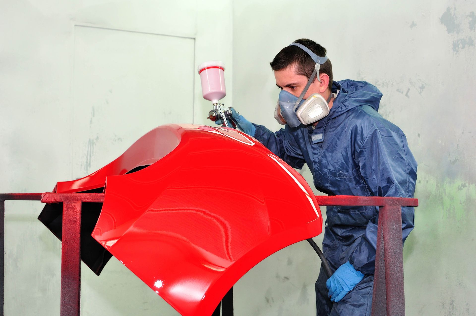 Person in a blue protective suit spray-painting a bright red car panel in a workshop
