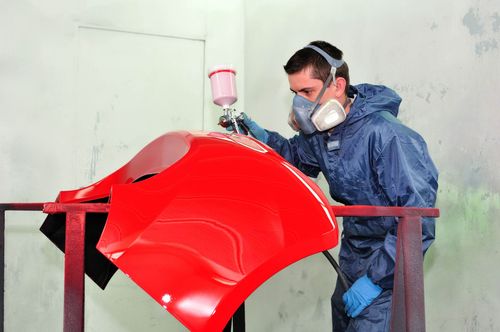 Person in a blue protective suit spray-painting a bright red car panel in a workshop