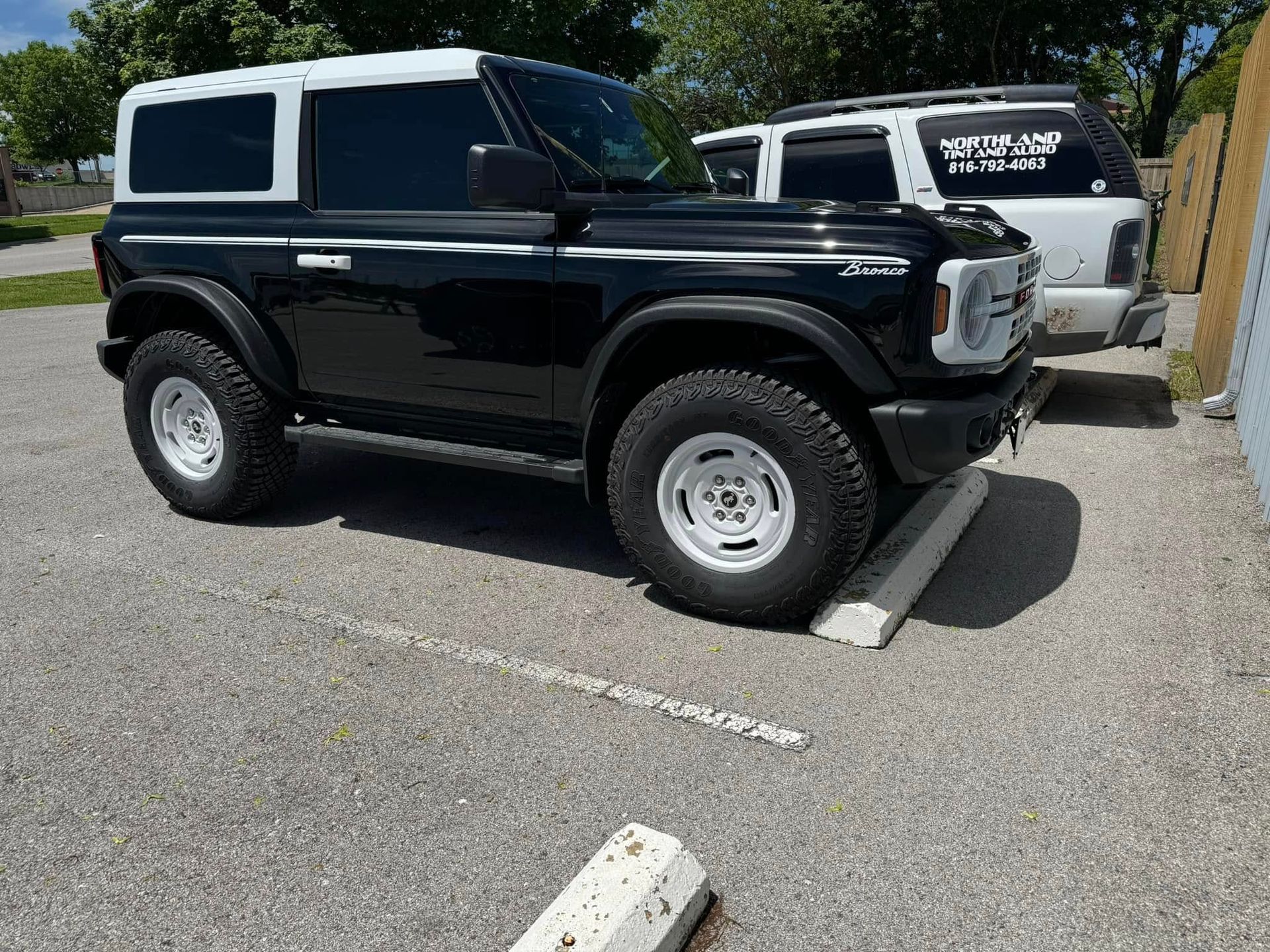 A black two-door Ford Bronco with a white hardtop and white wheels parked on an asphalt lot.