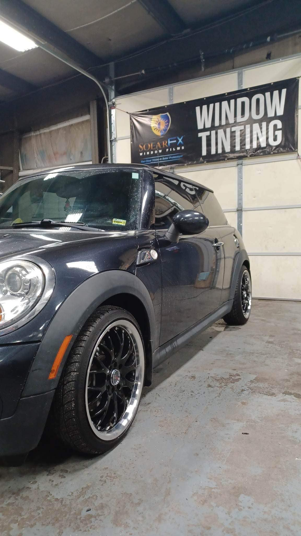 A black mini cooper is parked in a garage next to a window tinting sign.